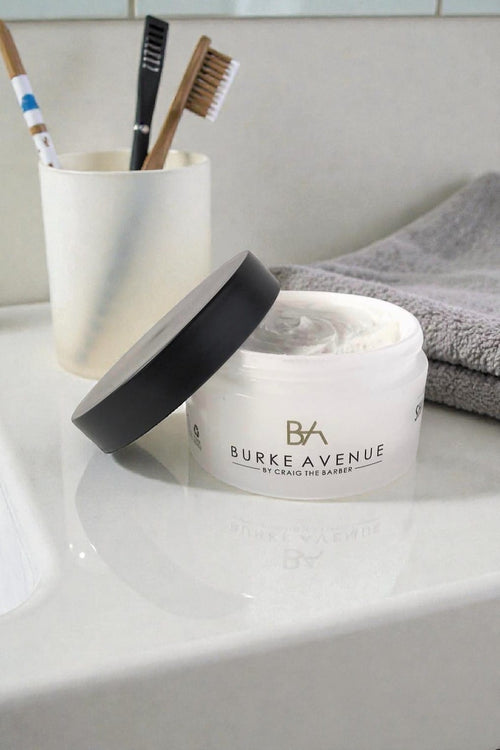 Open jar of Burke Avenue Moisturizing Shave Cream on bathroom counter beside toothbrushes and towel